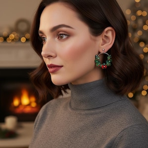 May include: A woman wearing a gray turtleneck sweater and hoop earrings. The earrings are decorated with green beads and small red accents, resembling miniature wreaths. The background features a blurred fireplace and Christmas lights.