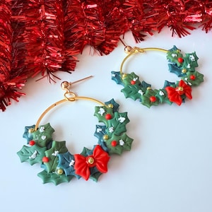 May include: A pair of gold hoop earrings with a festive Christmas theme. Each earring features a wreath design with green holly leaves, red berries, and a red bow. The earrings are set against a white background with red tinsel.
