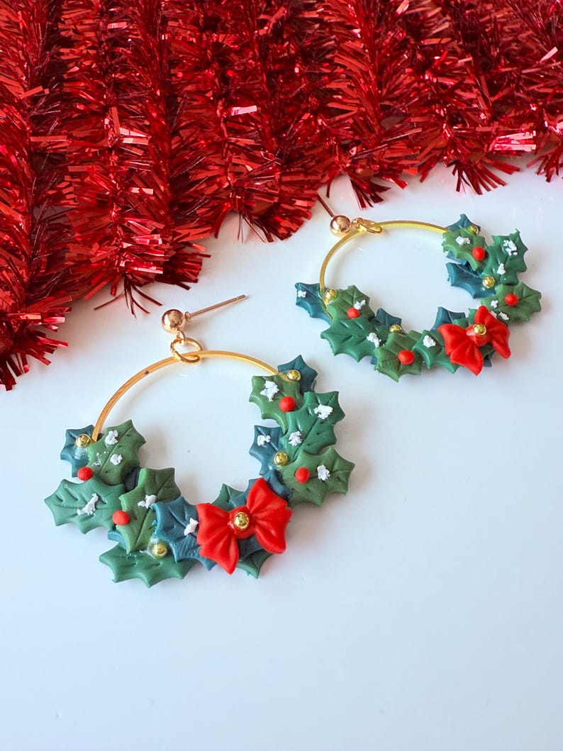 May include: A pair of gold hoop earrings with a festive Christmas theme. Each earring features a wreath design with green holly leaves, red berries, and a red bow. The earrings are set against a white background with red tinsel.