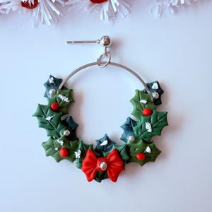 May include: A festive Christmas earring featuring a silver hoop adorned with a holly wreath design. The wreath includes green leaves, red berries, and a red bow with a silver center. The earring is set against a white background with white and red decorative elements.