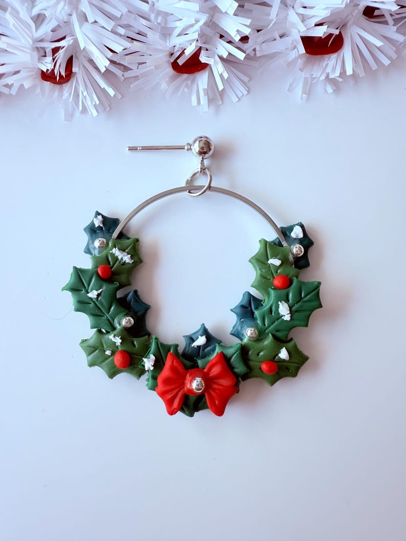May include: A festive Christmas earring featuring a silver hoop adorned with a holly wreath design. The wreath includes green leaves, red berries, and a red bow with a silver center. The earring is set against a white background with white and red decorative elements.