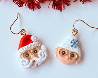 May include: A pair of dangle earrings featuring sculpted clay faces of Santa Claus and a character in a white winter hat. Santa wears a red hat with white trim and gold wire glasses. The other character wears a white hat with a blue snowflake.