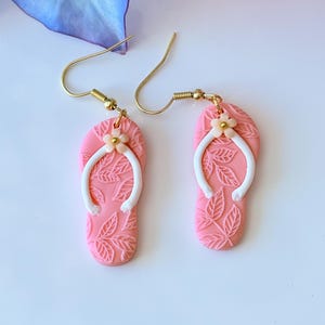 Könnte beinhalten: Rosa Flip-Flop-Ohrringe mit weißen Riemen und einem kleinen Blumen-Detail. Die Ohrringe sind aus Polymer Clay gefertigt und haben goldene Haken.