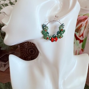 May include: A silver hoop earring featuring a miniature Christmas wreath design. The wreath is composed of green holly leaves, red berries, and a small red bow. The earring is displayed on a white mannequin head.
