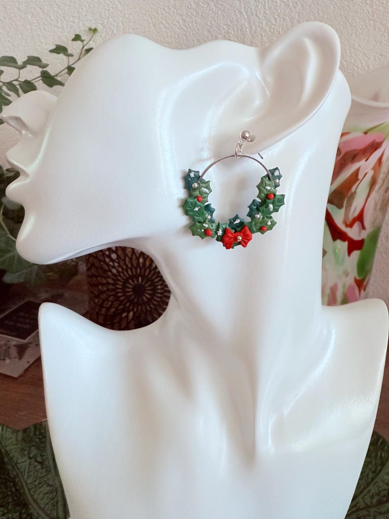 May include: A silver hoop earring featuring a miniature Christmas wreath design. The wreath is composed of green holly leaves, red berries, and a small red bow. The earring is displayed on a white mannequin head.