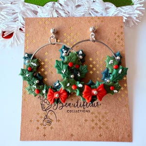 May include: Silver hoop earrings with a festive Christmas theme. Each earring features a green holly wreath design with red berries, red bows, and silver accents. The earrings are displayed on a brown card with the text "Beautiful Collections".