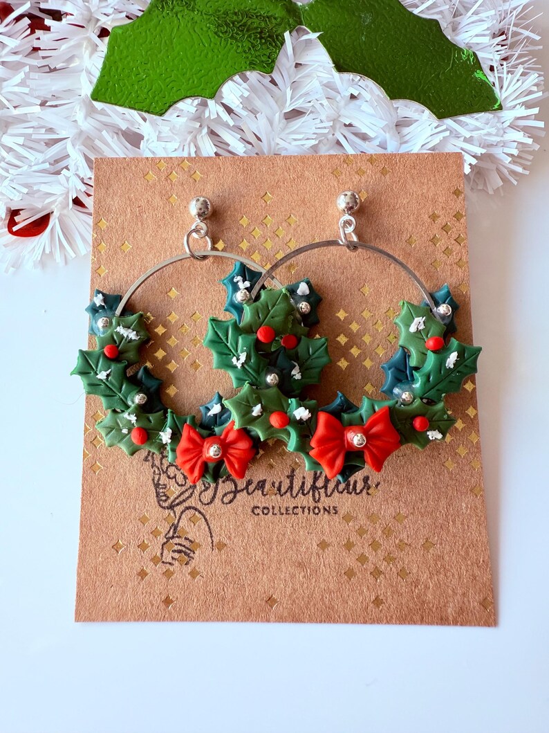 May include: Silver hoop earrings with a festive Christmas theme. Each earring features a green holly wreath design with red berries, red bows, and silver accents. The earrings are displayed on a brown card with the text "Beautiful Collections".