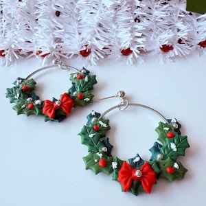 May include: A pair of Christmas-themed hoop earrings. Each earring features a green holly wreath design with red berries and a red bow. The earrings have silver-colored hoops and are set against a white background with decorative garland.