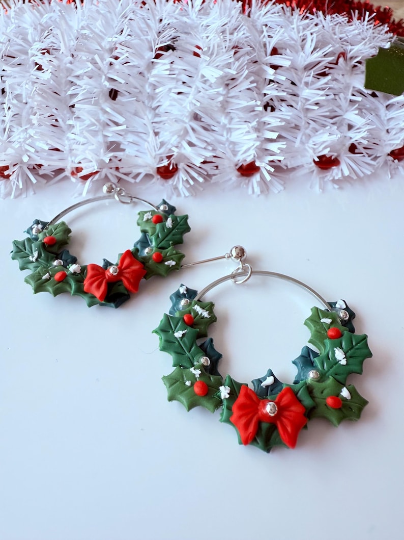 May include: A pair of Christmas-themed hoop earrings. Each earring features a green holly wreath design with red berries and a red bow. The earrings have silver-colored hoops and are set against a white background with decorative garland.