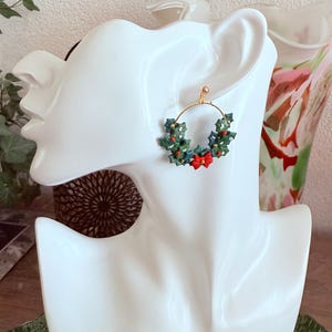 May include: A gold hoop earring featuring a festive Christmas wreath design. The wreath is composed of green leaves, red berries, and a red bow. The earring is displayed on a white mannequin head, showcasing its details and design.