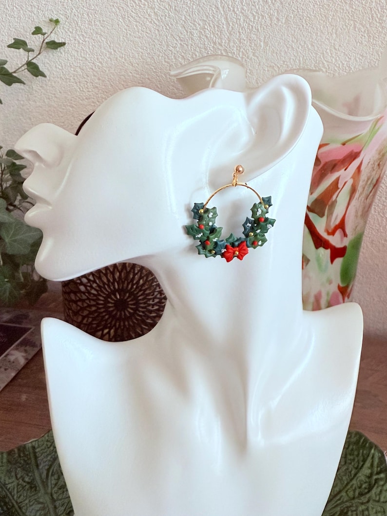 May include: A gold hoop earring featuring a festive Christmas wreath design. The wreath is composed of green leaves, red berries, and a red bow. The earring is displayed on a white mannequin head, showcasing its details and design.