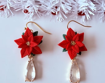 May include: A pair of dangle earrings featuring red poinsettia flowers with green leaves and gold-tone centers, suspended from gold-tone hooks with clear, teardrop-shaped faceted crystals hanging below each flower.