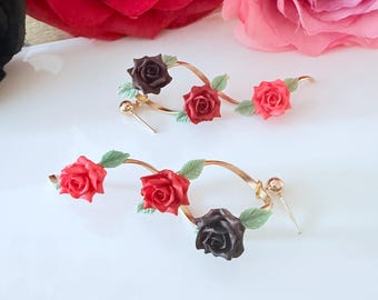 May include: A pair of gold-tone hoop earrings featuring three-dimensional rose blossoms in shades of dark brown, bright red, and light pink. Each rose is accented with small green leaves attached to the curved gold metal wire frame.