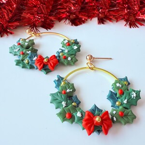 May include: A pair of gold hoop earrings featuring a Christmas wreath design. The wreaths are made of green and blue leaves, red berries, and a red bow. The earrings are festive and suitable for the holiday season.