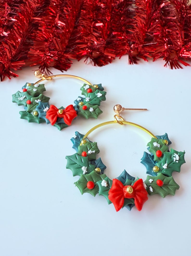 May include: A pair of gold hoop earrings featuring a Christmas wreath design. The wreaths are made of green and blue leaves, red berries, and a red bow. The earrings are festive and suitable for the holiday season.
