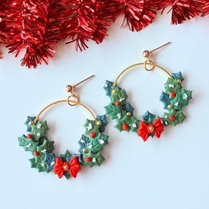 May include: A pair of festive hoop earrings. Each earring features a gold-colored hoop adorned with green and blue holly leaves, red berries, and a red bow. The earrings are designed to resemble miniature Christmas wreaths.