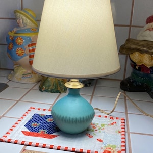 May include: A turquoise ceramic table lamp with a white fabric lampshade. The lamp is sitting on a red and white patchwork quilt.