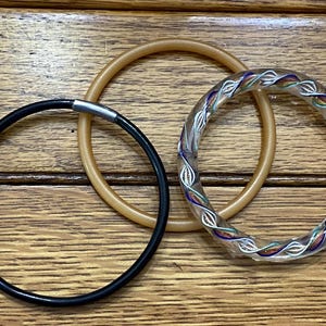 May include: Three decorative bracelets. One is black with a silver clasp, another is a light brown, and the third is clear with a colorful, intricate design. The bracelets are circular and rest on a wooden surface.