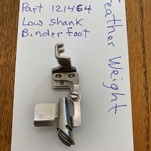 May include: Silver low shank binder foot sewing machine attachment. The part number is 121464. The text "Feather Weight" is written on the paper.