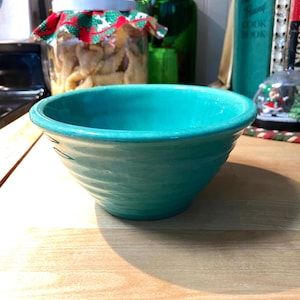 May include: A teal blue ceramic mixing bowl with a white interior. The bowl has a slightly textured surface and is sitting on a wooden surface.