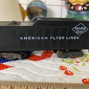 May include: Black toy train engine with the words "American Flyer Lines" and "Reading Lines" printed on the side. The train is sitting on a white and red quilted fabric.
