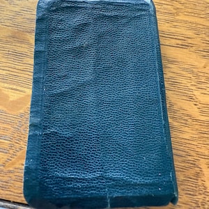 May include: A closed, rectangular book with a dark teal leather cover. The cover has a textured, pebbled appearance. The edges of the book are slightly worn, and the spine is visible. The book is resting on a wooden surface.