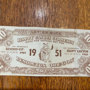May include: A vintage Happy Canyon Currency ticket from the 1951 Round-Up in Pendleton, Oregon. The ticket is brown and white with a drawing of a cowboy riding a bucking bronco. The ticket is worth 10 "bucks" and is not redeemable.
