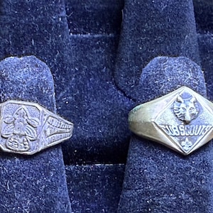 May include: Two vintage Cub Scout rings. One ring features a fleur-de-lis emblem, and the other has a wolf's head and the words "Cub Scouts". Both rings are made of a tarnished metal and are displayed on a dark blue velvet surface.