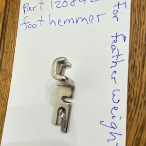 May include: Silver sewing machine foot for a featherweight foot hemmer. The part number is 12084/2.  The text "Siman Co" is written on the card.