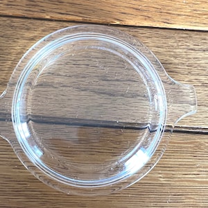 May include: A clear, round glass lid with two small handles. The lid has a slightly raised, concentric ring design. The object is likely a replacement lid for a casserole dish or similar cookware.