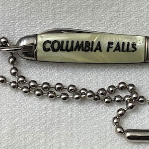 May include: A white pocket knife with a silver chain and a black inscription that reads "COLUMBIA FALLS".