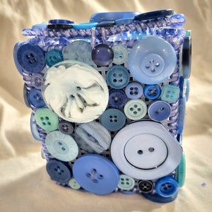 May include: A blue and white decorative box made of buttons. The box is covered in a variety of blue and white buttons, including some with a floral design. The box is made of a clear plastic material and has a woven pattern.