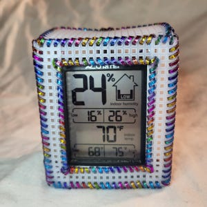 May include: A white digital indoor humidity and temperature gauge with a black display and a black frame. The gauge is surrounded by a white plastic frame with a colorful beaded border. The display shows the current humidity is 24% and the temperature is 70 degrees Fahrenheit.