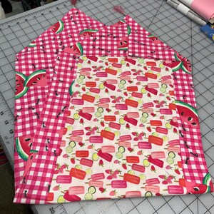 May include: Two handmade fabric placemats. One features a pink and white gingham pattern with watermelon slices. The other has a white background with colorful popsicle and fruit designs. Both have pink tassels.
