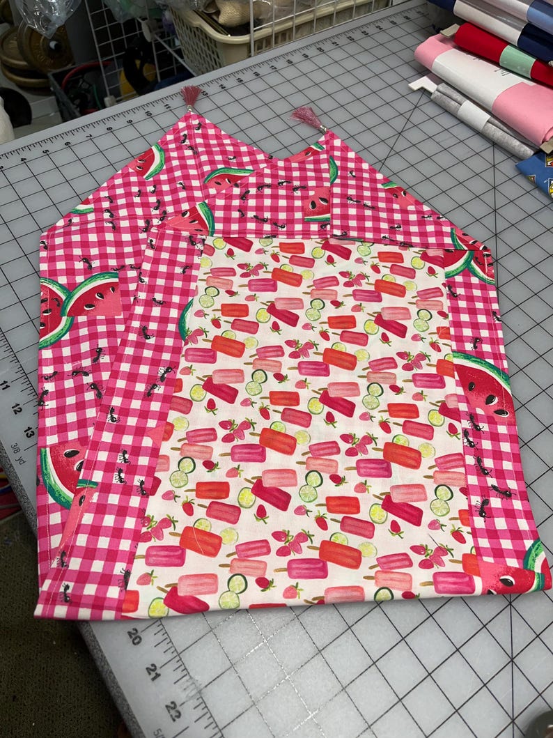 May include: Two handmade fabric placemats. One features a pink and white gingham pattern with watermelon slices. The other has a white background with colorful popsicle and fruit designs. Both have pink tassels.