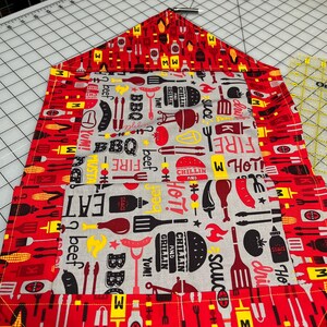 May include: A red and gray BBQ-themed table runner with a repeating pattern of grilling tools, food items, and text. The central panel features words like "BBQ," "Eat," and "Grillin'" alongside images of burgers, sausages, and sauces.