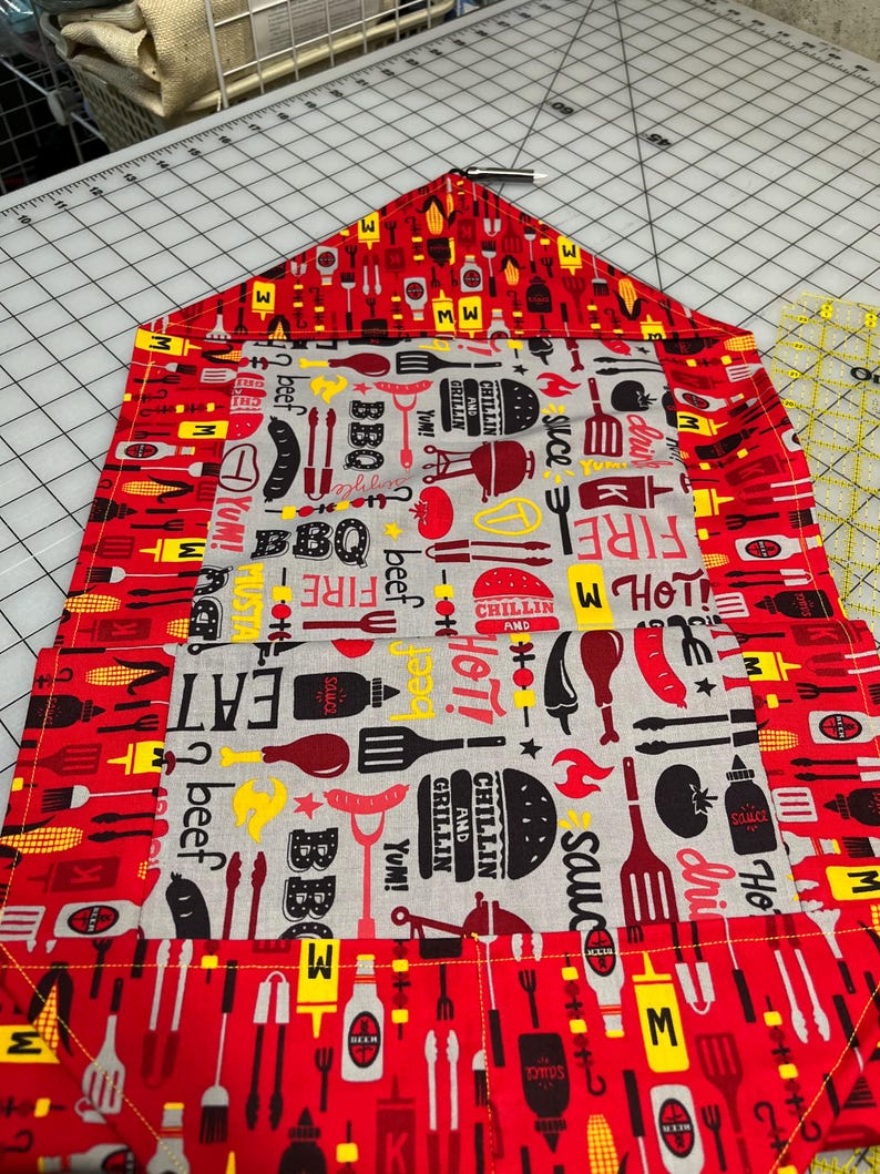 May include: A red and gray BBQ-themed table runner with a repeating pattern of grilling tools, food items, and text. The central panel features words like "BBQ," "Eat," and "Grillin'" alongside images of burgers, sausages, and sauces.