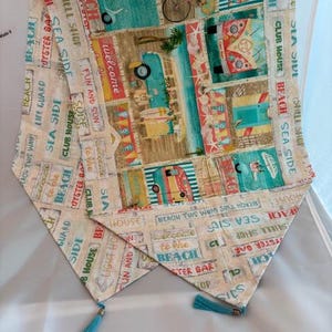 May include: A decorative table runner with a beach theme. The fabric features colorful illustrations of beach scenes, vintage campers, and text like "Welcome to the Beach." It has turquoise tassels at the ends.