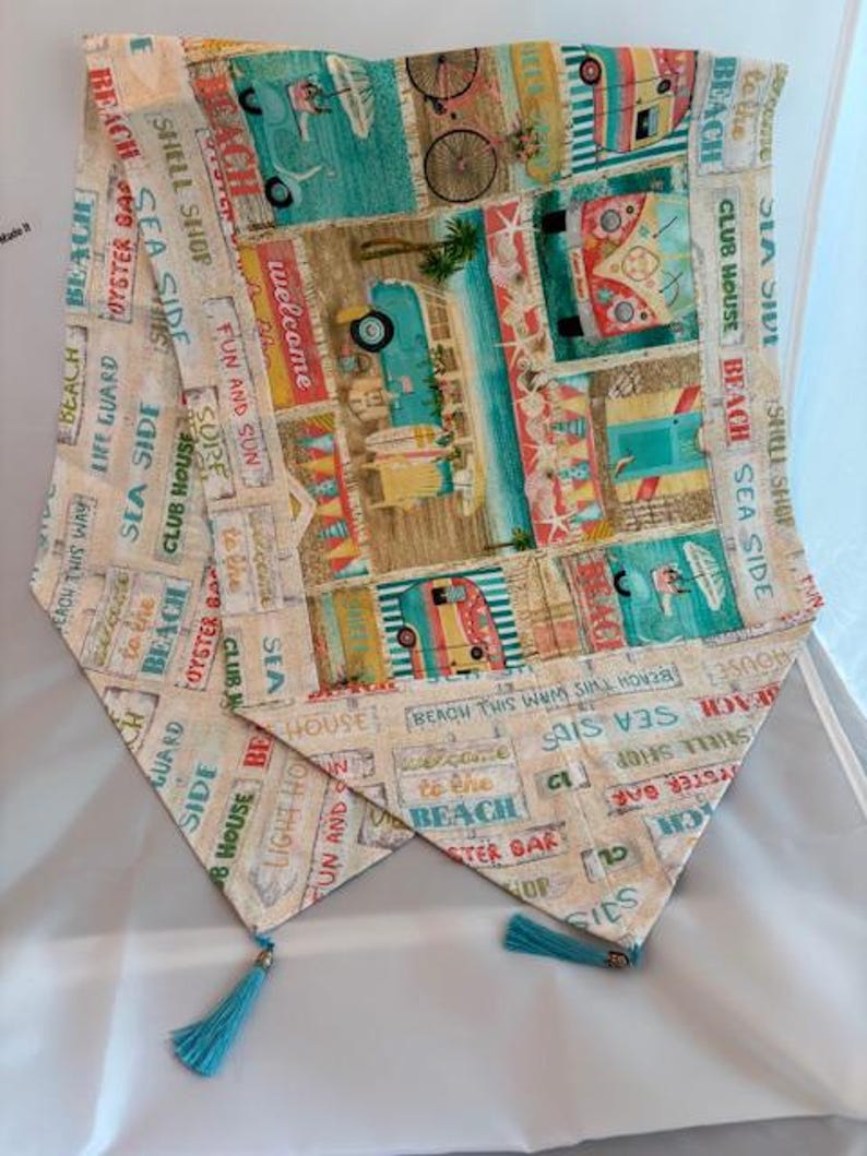 May include: A decorative table runner with a beach theme. The fabric features colorful illustrations of beach scenes, vintage campers, and text like "Welcome to the Beach." It has turquoise tassels at the ends.