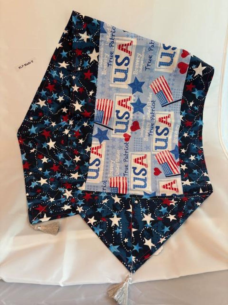 May include: A patriotic table runner with a blue, red, and white color scheme. The runner features star and flag patterns, with the word "USA" prominently displayed. Tassels adorn the ends. The design evokes a sense of American pride and celebration.