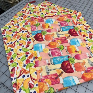 May include: A stack of colorful fabric placemats. One placemat features a pattern of various cocktails, while the others display a vibrant fruit design with peaches, strawberries, and blueberries. The placemats have a rectangular shape.