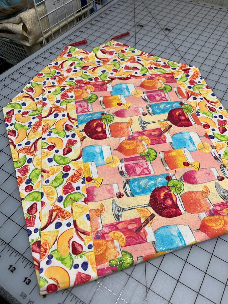May include: A stack of colorful fabric placemats. One placemat features a pattern of various cocktails, while the others display a vibrant fruit design with peaches, strawberries, and blueberries. The placemats have a rectangular shape.