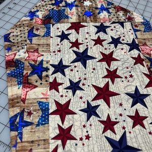 May include: A patriotic-themed table runner featuring a repeating pattern of red, white, and blue stars on a cream background. The runner has a wood grain pattern on the edges and is likely made of fabric.