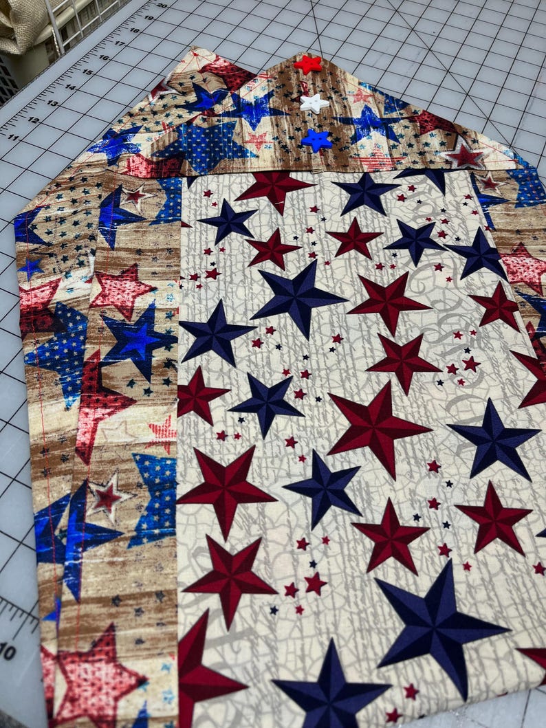 May include: A patriotic-themed table runner featuring a repeating pattern of red, white, and blue stars on a cream background. The runner has a wood grain pattern on the edges and is likely made of fabric.