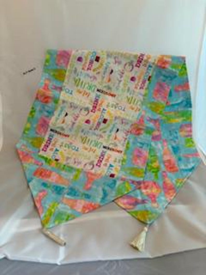 May include: A colorful table runner with a light blue background and a central panel featuring text and images related to drinks and celebrations. The runner has a length of approximately 60 inches and includes tassels at each end.