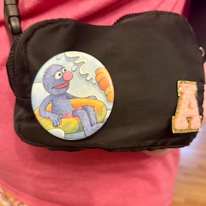 May include: Black nylon crossbody bag with a round Grover button and a gold glitter "A" patch. The button features Grover from Sesame Street in a yellow inner tube. The bag has a black strap and a zipper closure.