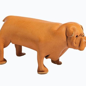 May include: A tan leather bulldog figurine. The sculpture shows the dog's face, body, and legs in detail. The bulldog is standing on all fours, with a realistic texture and colour. The figurine is made of leather.