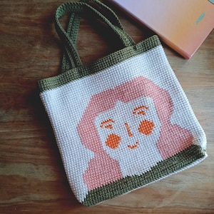 Puede incluir: Bolso tote hecho a mano con un diseño de cara pixelada en rosa y naranja sobre fondo blanco. El bolso tiene ribetes y asas verdes. Se muestra sobre una superficie de madera.