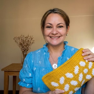 Puede incluir: Bolso de mano amarillo mostaza con lunares blancos, de forma rectangular y tejido a ganchillo. La persona que lo sostiene lleva una blusa azul y sonríe. El fondo es neutro.