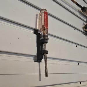 Puede incluir: Un destornillador con mango transparente y etiqueta roja está montado en una pared de listones blancos. Una caña de pescar y un carrete también están montados en la pared, lo que sugiere un sistema de almacenamiento.